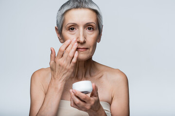 mature woman with grey hair holding container and applying cosmetic cream isolated on grey
