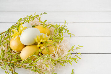 Easter yellow eggs in a decorative nest decorated with bright spring flowers and willow green branches on a white table background, top view, copy space. Festive decor, easter card