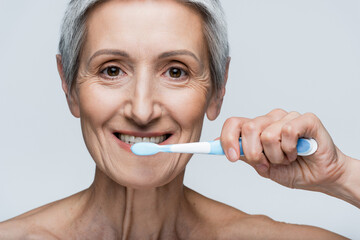 happy middle aged woman holding toothbrush and brushing teeth isolated on grey