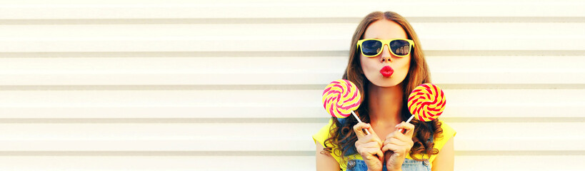 Summer portrait of beautiful young woman blowing her lips with red lipstick holding colorful lollipop on stick over white background