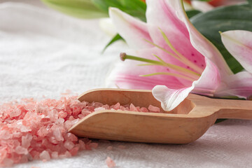 Pink salt crystals in a wooden scoop and beautiful lily flower on white background. Spa set. Floral spa background. Selective focus, copy space
