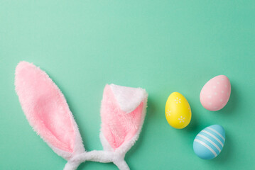 Top view photo of easter decorations bunny ears headband pink yellow and blue easter eggs on isolated turquoise background with copyspace