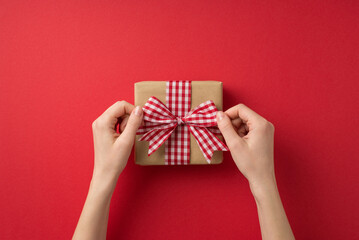 First person top view photo of valentine's day decorations woman's hands tying checkered ribbon bow on craft paper giftbox on isolated red background with copyspace