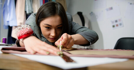 Beautiful asian woman fashion designer measuring and making outlines of clothing sewing pattern on fabric. Young female tailor working at atelier.