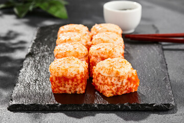 Baked Maki sushi on dark slate. Hot california maki with tobiko. Sushi roll with masago caviar, cheese topped baked oven. Style concept japanese menu with black background, leaves and hard shadow.