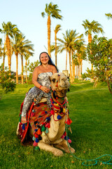 A brunette woman in a cape on her head firmly clings to the lying camel on the grass against the backdrop of palm trees.