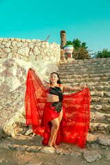 A woman in a red suit dances an oriental dance on a stone staircase against a blue sky.