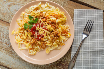 Pasta bolognese in tomato garnished with herbs and cheese in a plate on a wooden table next to a fork and a napkin.