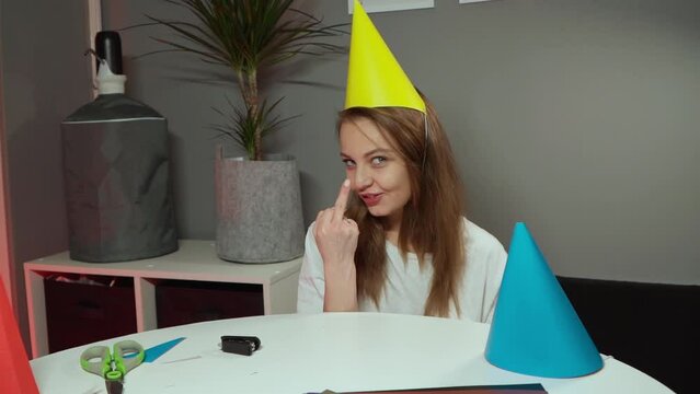 Caucasian girl sits in a whate T-shirt and smiles. A European woman in birthday cap showing middle finger. Decor for the holiday with your own hands. Homemade attributes for a birthday. Gray walls