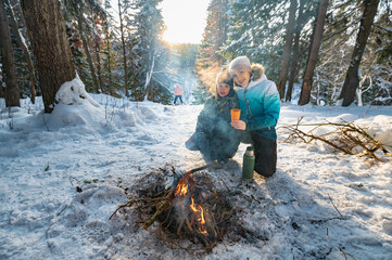A woman and a boy at a campfire in winter in the forest drink tea from a thermos. - 488838681