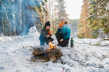 A woman and a boy at a campfire in winter in the forest drink tea from a thermos.