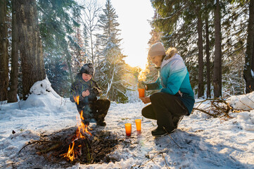 A woman and a boy at a campfire in winter in the forest drink tea from a thermos. - 488838684
