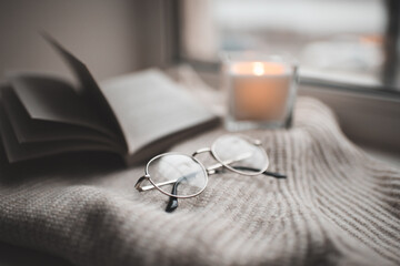 Fototapeta na wymiar Scented candle stay on open paper book with glasses on knit cloth sweater closeup at home. Cozy hygge atmosphere. Aromatherapy. 