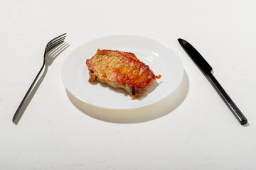 A piece of fried meat on a white plate. Black knife and fork. Bad fatty food. White background. Diet keto concept.