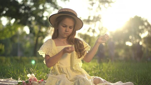 Wide shot thoughtful cute girl sitting at golden sunset in spring summer park. Beautiful Caucasian child in yellow dress and straw hat playing with grass resting on meadow in sunbeam outdoors