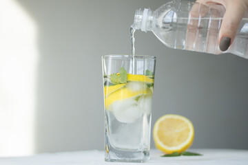 woman pouring water from bottle into glass in the room. a female hand pours water from a bottle into a glass with ice and lemon, mint on a gray background