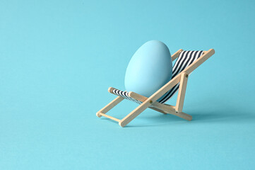 Creative funny composition with Easter egg while sitting on deck chair on pastel blue background.