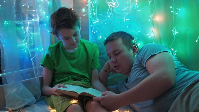 dad and son are reading a book lying on the couch. into the light of the garlands. bedroom decoration. a beautiful holiday