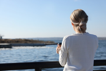 Fototapeta na wymiar Lonely woman with cup of drink near river on sunny day, back view