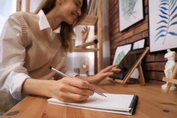 Young woman using tablet while drawing in sketchbook with pencil at wooden table indoors, focus on hand
