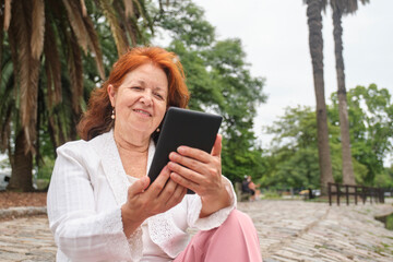 Mature hispanic woman reading an ebook on an electronic reader sitting in a park