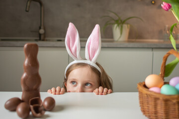 Cute little girl wearing bunny ears eating chocolate Easter rabbit. Kid playing egg hunt on Easter....