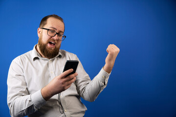 Successful bearded man in a casual shirt and glasses on a blue background is very happy and excited. Jackpot winner with hands up, smiling and shouting about success. Gesture of the winner.