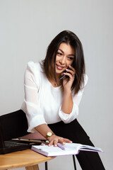 business woman posing while working at her desk. A laptop and some papers were placed in front of her. She is dressed in a strict black suit and talking on the phone. Isolated, white background.