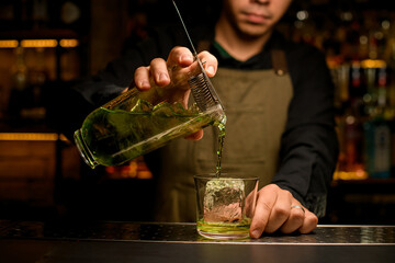 hand of barman gently holds mixing cup with cold drink and pour it into a glass with ice cube