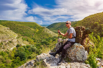 A hiker with a backpack rests sitting on a stone while descending from the mountains into a green...