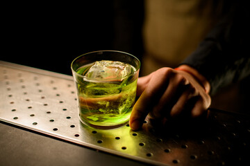 Cocktail glass drink with ice cube stands on the bar counter and hand of male barman holds it