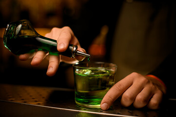 Selective focus of bottle with green essences, a drop of which adds to glass with a cocktail