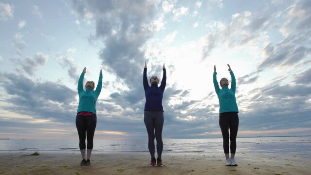 Group of three mature women practicing yoga at sunset. Women doing standing backbend and side bend poses with raised arms standing near sea under cloudy evening sky, instructor explaining exercise