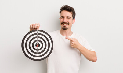 young handsome man smiling cheerfully, feeling happy and pointing to the side. dart target concept - obrazy, fototapety, plakaty
