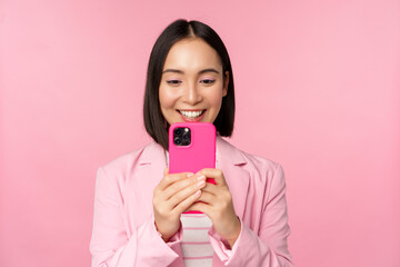 Image of smiling asian corporate woman in suit looking, watching on smartphone app, using mobile...