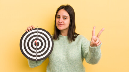 young hispanic woman smiling and looking happy, gesturing victory or peace. darts target concept