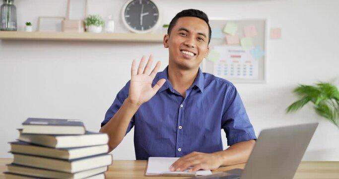 Portrait of young Asian man in blue shirt smiling and waving hand while sitting at workdesk. Male looking at camera.