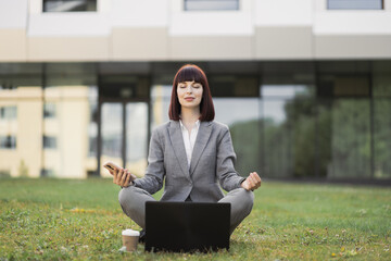 Pretty young female executive practicing yoga, sitting with eyes closed outside office on green...