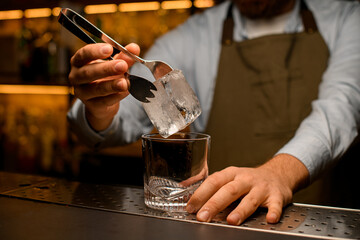 selective focus on square piece of ice held by the male hand with tongs over glass