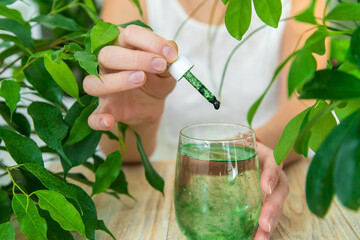 Woman dripping chlorophyll supplement into a glass of water. Selective focus. - obrazy, fototapety, plakaty