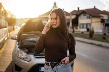 Fototapeta na wymiar One young adult caucasian woman standing by her vehicle with open hood broken failed engine holding a phone calling towing service for help on the road Roadside assistance concept autumn or spring day