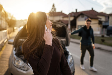Fototapeta premium One young adult caucasian woman standing by her vehicle with open hood broken failed engine holding a phone calling towing service for help on the road Roadside assistance concept autumn or spring day
