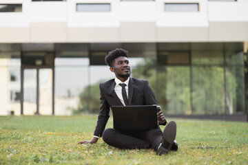 Approval of the project. Hardworking cheerful African businessman sitting on green grass outside modern office, showing thumb up while using laptop. Successful business, technologies concept