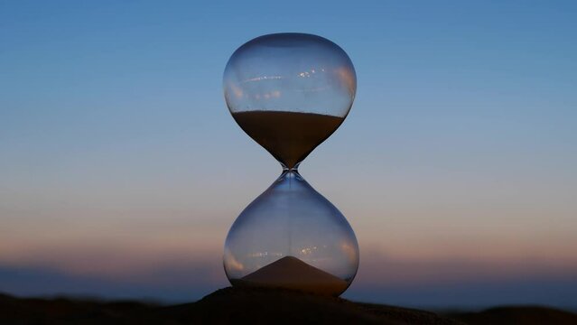 Hourglass time lapse shot, evening sky on background. Quickly night falls, picture becomes dark. Slowly at first, and then faster and quite quickly, remains of sand from upper bulb falls down