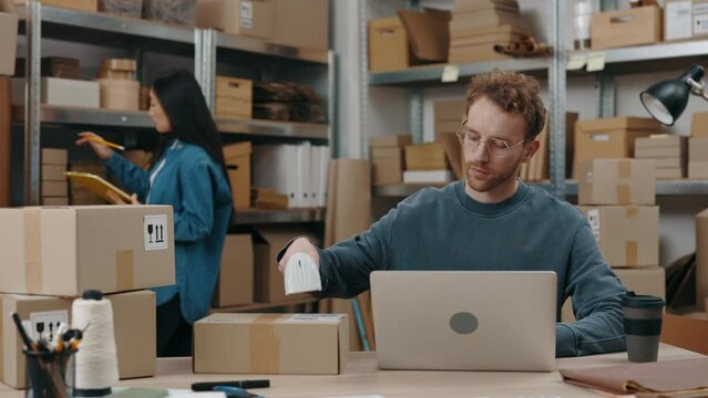 Attentive caucasian man wearing glasses sitting at the laptop and scanning parcels while his asian brunette colleague sorting through parcels. Small business concept.