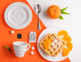 On a white-orange background, cups for tea, Belgian waffles in a plate, tangerine and sugar. Tea party concept.