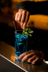 Clear glass of blue cold drink on bar and the bartender decorating it with fresh green mint leaves