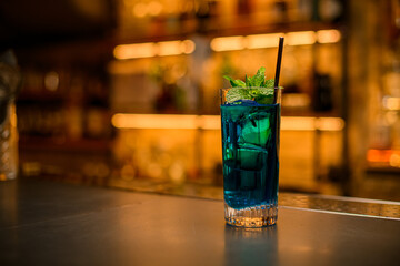 Beautiful view of glass of blue cold drink with steel drinking straw and green mint leaves on bar counter.