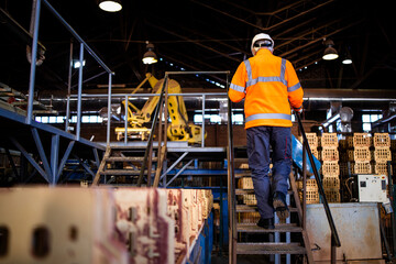 Factory worker in safety equipment climbing metal staircase to get to industrial production machine.