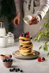 Female pouring honey maple syrup on traditional blueberry pancakes with raspberries and walnuts stacked on top of each other